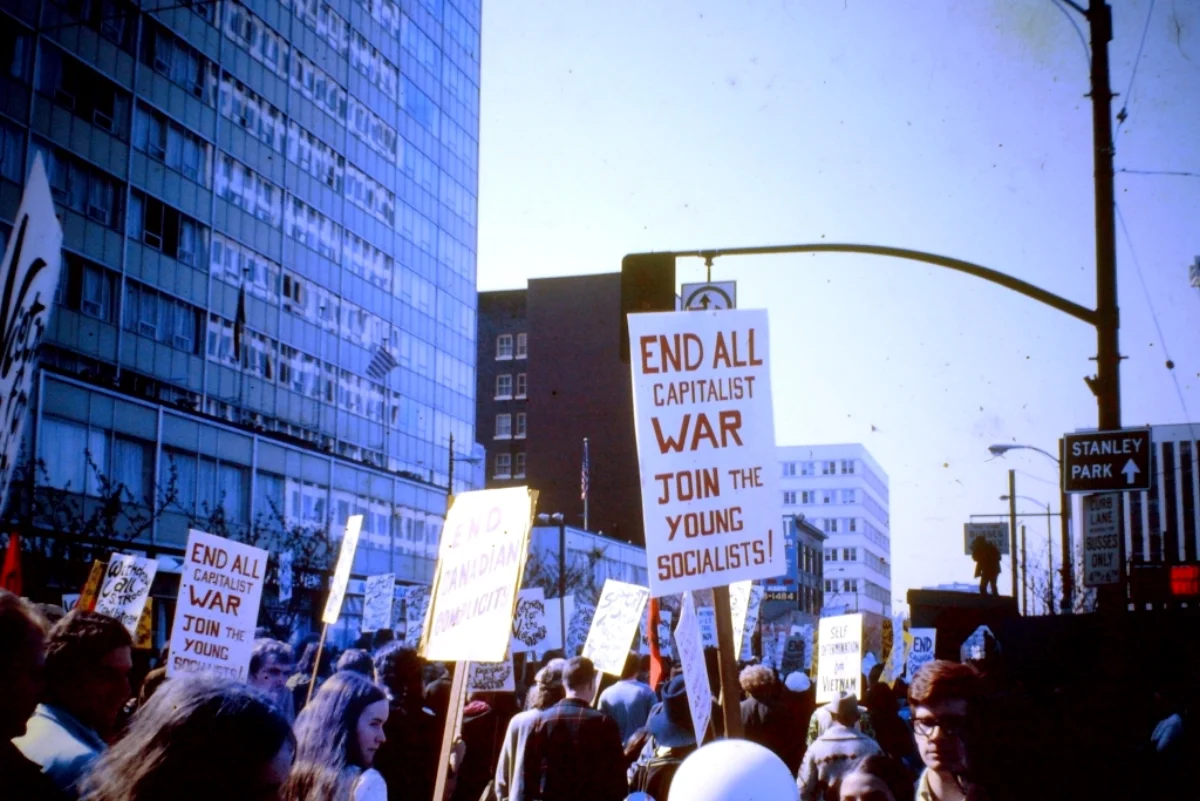 Color photograph from 1968 of a large crowd at an anti-Vietnam War demonstration in Vancouver, BC; protestors raise hand-lettered signs reading 'END ALL CAPITALIST WAR — JOIN THE YOUNG SOCIALISTS' and 'END CANADIAN COMPLICITY' against a backdrop of mid-century office buildings
