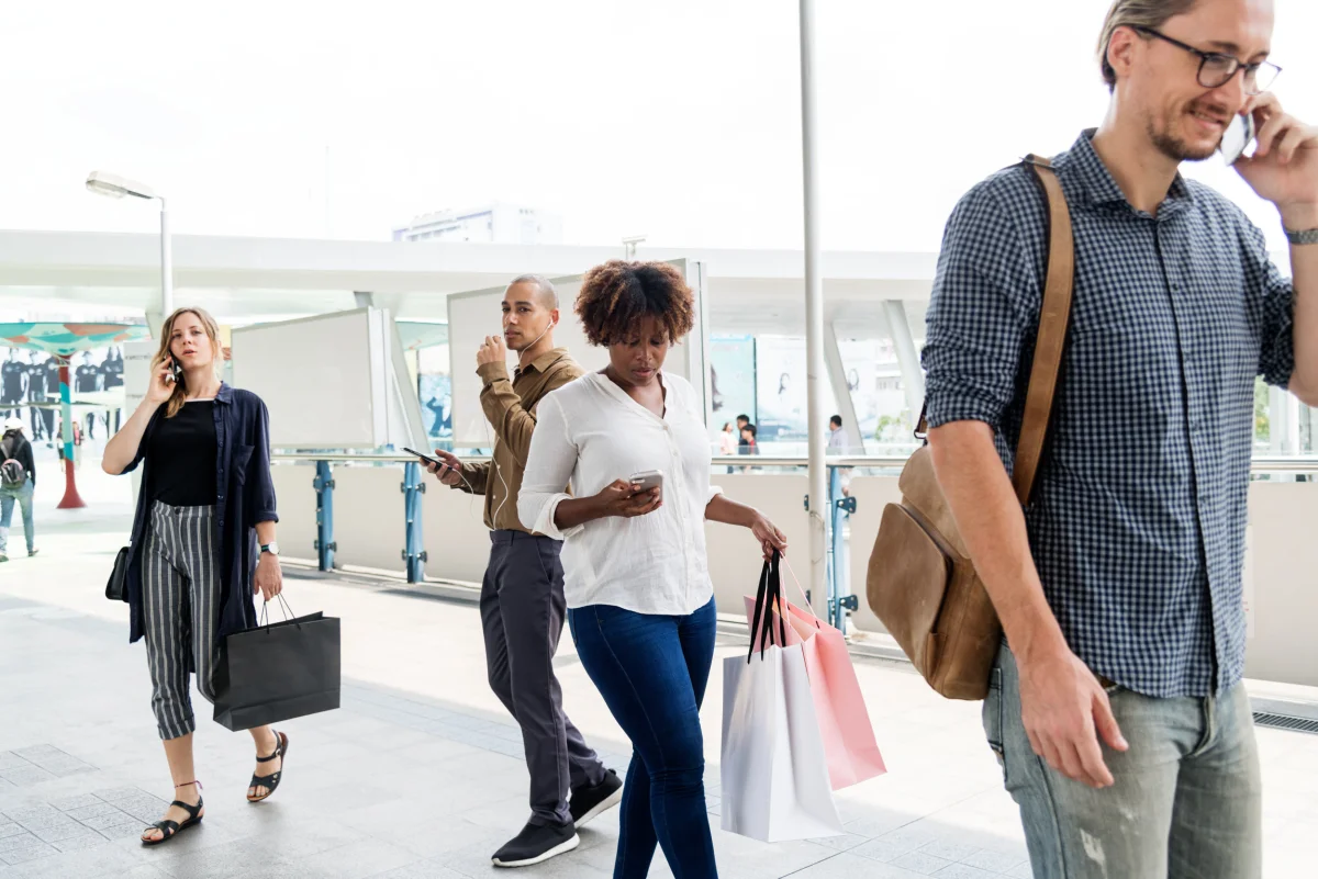 Color candid photograph of four pedestrians on an outdoor walkway, three of them looking down at smartphones or holding them to their ears, the fourth talking on a mobile phone
