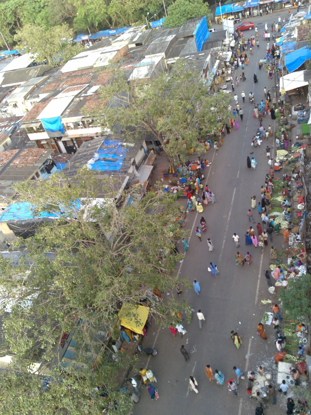 Aerial color photograph looking down on Dharavi, Mumbai: a dense, low-rise informal settlement of corrugated-tin and tarp-covered buildings packed tightly along narrow alleys with crowds of pedestrians flowing through a market street between rows of shacks