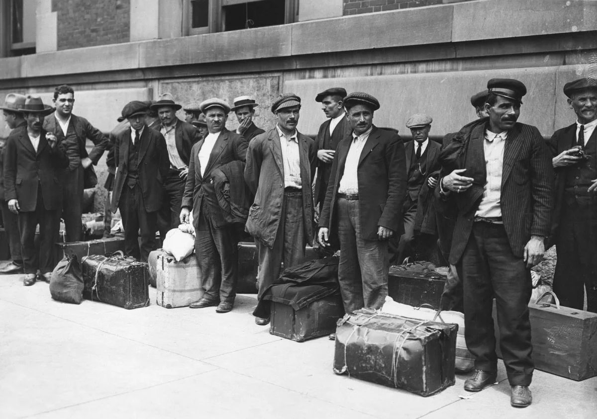 Black-and-white historical photograph of about a dozen Italian immigrant men in flat caps and three-piece suits standing with battered suitcases and bundled belongings against a stone wall at Ellis Island in the early 20th century