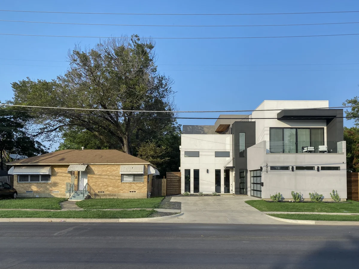 Color photograph of two adjacent houses on the same street in Old East Dallas: on the left, a small single-story bungalow with peeling paint; on the right, a brand-new three-story modern home in white stucco and glass that towers over the older structure