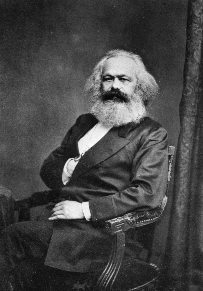 Black-and-white seated portrait photograph of Karl Marx in his sixties, with a full white beard and unruly white hair, wearing a dark three-piece suit with a watch chain visible across his vest