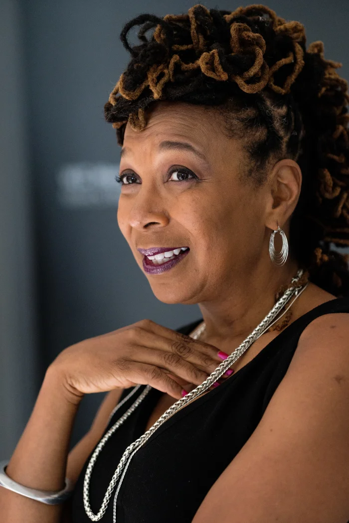 Color portrait photograph of legal scholar Kimberlé Crenshaw, a Black woman with locs styled in a high updo, wearing a black sleeveless top with silver chain necklaces and a silver bangle, smiling slightly while resting one hand near her collarbone