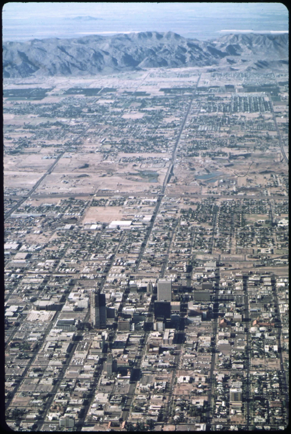 Aerial color photograph from 1972 showing downtown Phoenix in the foreground giving way to a vast grid of low-density suburban tract housing extending to the horizon, with bare desert and distant mountains beyond