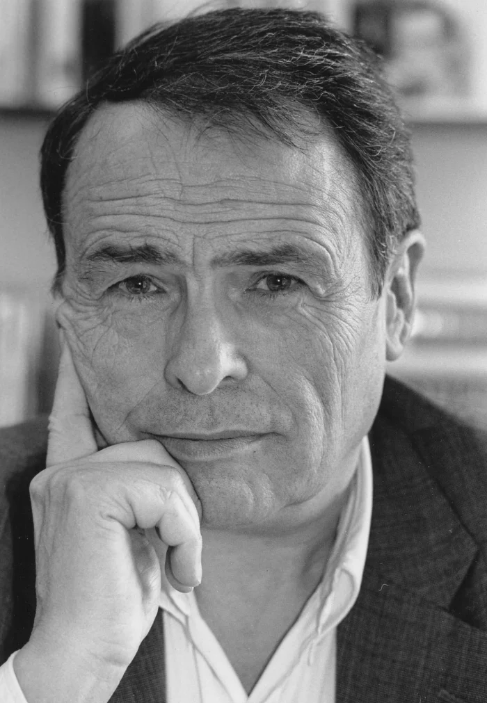 Black-and-white close-up portrait of French sociologist Pierre Bourdieu in middle age, resting his chin on his hand and looking thoughtfully into the camera, in a study with bookshelves visible behind him