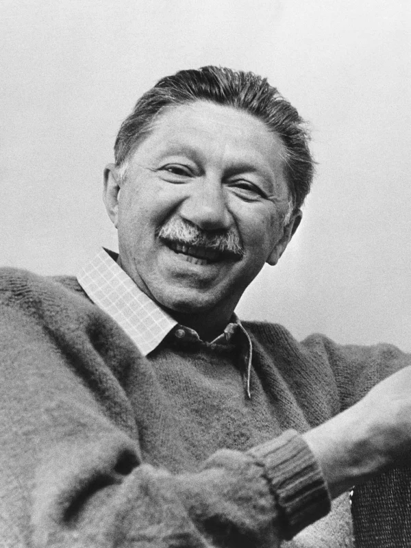 Black-and-white portrait of Abraham Maslow, a middle-aged man with a mustache wearing a cardigan over a button-down shirt, smiling broadly toward the camera
