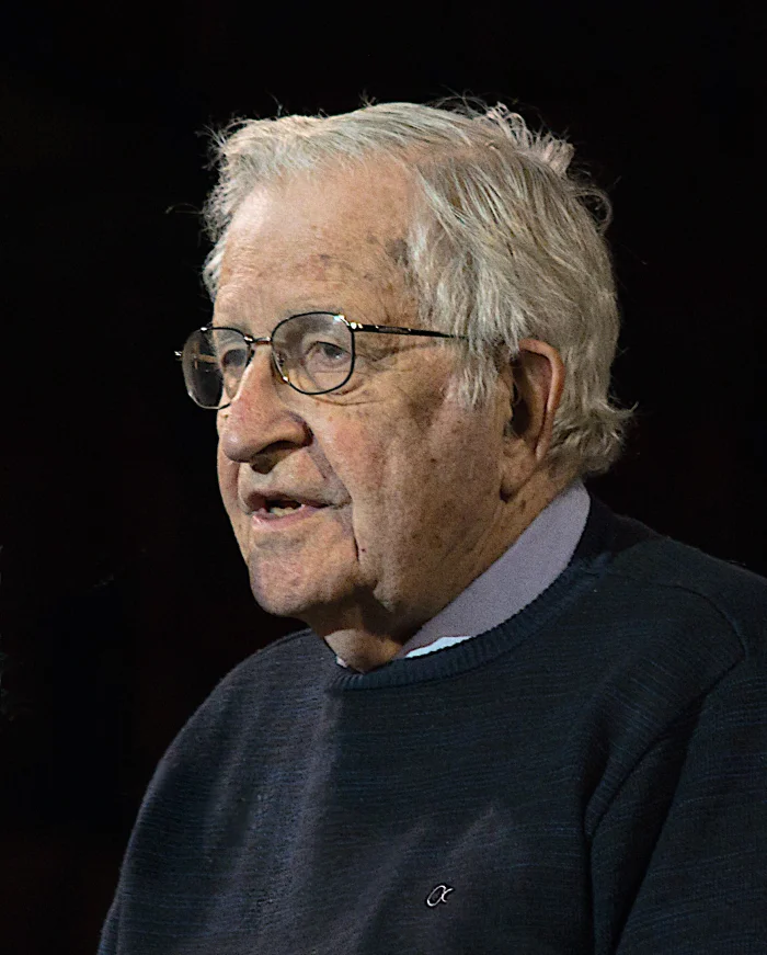 Color portrait of Noam Chomsky in his late eighties, white hair and beard, looking directly at the camera against a dark background