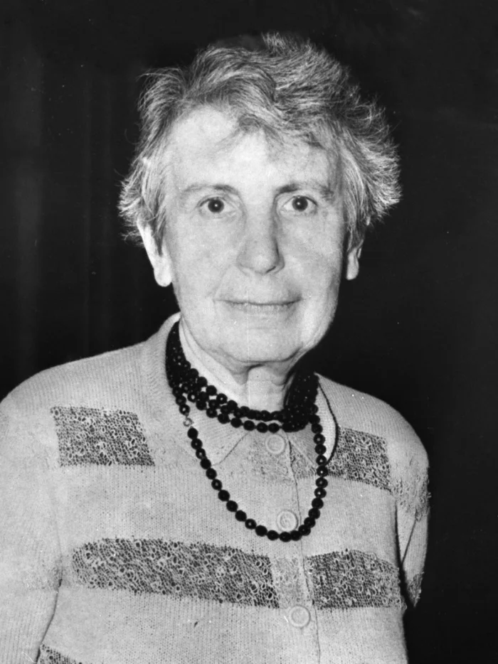 Black-and-white portrait of an older Anna Freud wearing a knit cardigan and a long string of dark beads, looking directly at the camera