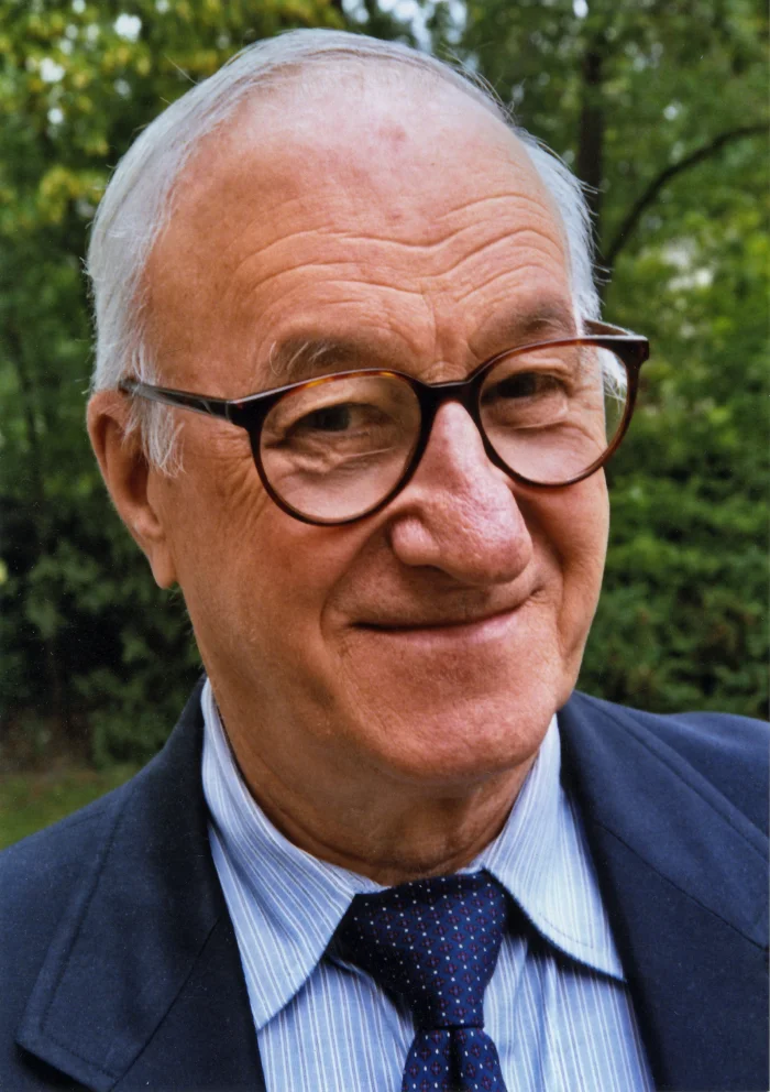 Color portrait photograph of Albert Bandura, an older man with white hair and large glasses, smiling and wearing a dark blue suit and patterned tie