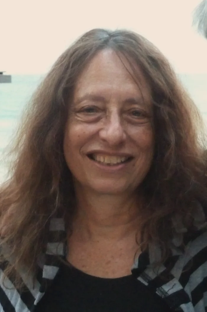 Color photograph of Carol Gilligan, a woman with long curly brown hair, smiling broadly at the camera against an outdoor seaside background