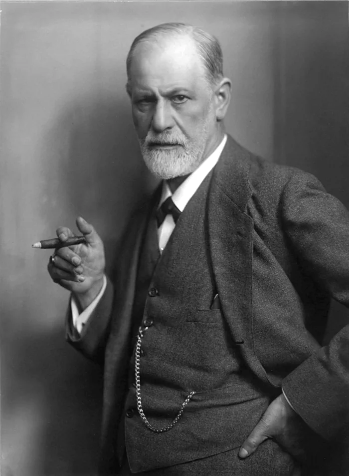 Black-and-white studio portrait of Sigmund Freud in a three-piece suit, holding a cigar in his right hand, looking sternly at the camera