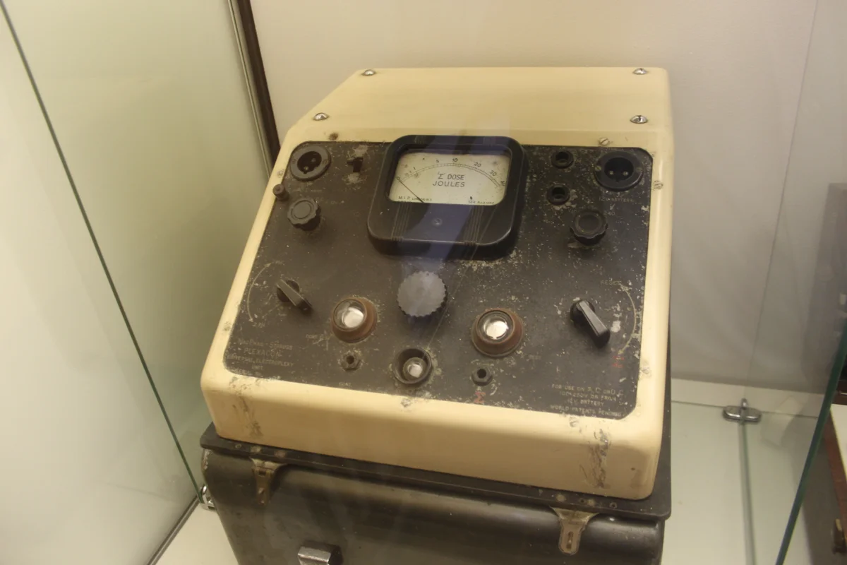 A vintage cream-colored Siemens Konvulsator III electroconvulsive therapy machine sitting on a wooden display surface, with a large central dial showing seconds, several smaller knobs and switches labeled in German, and two black handheld electrode paddles connected by cables in front