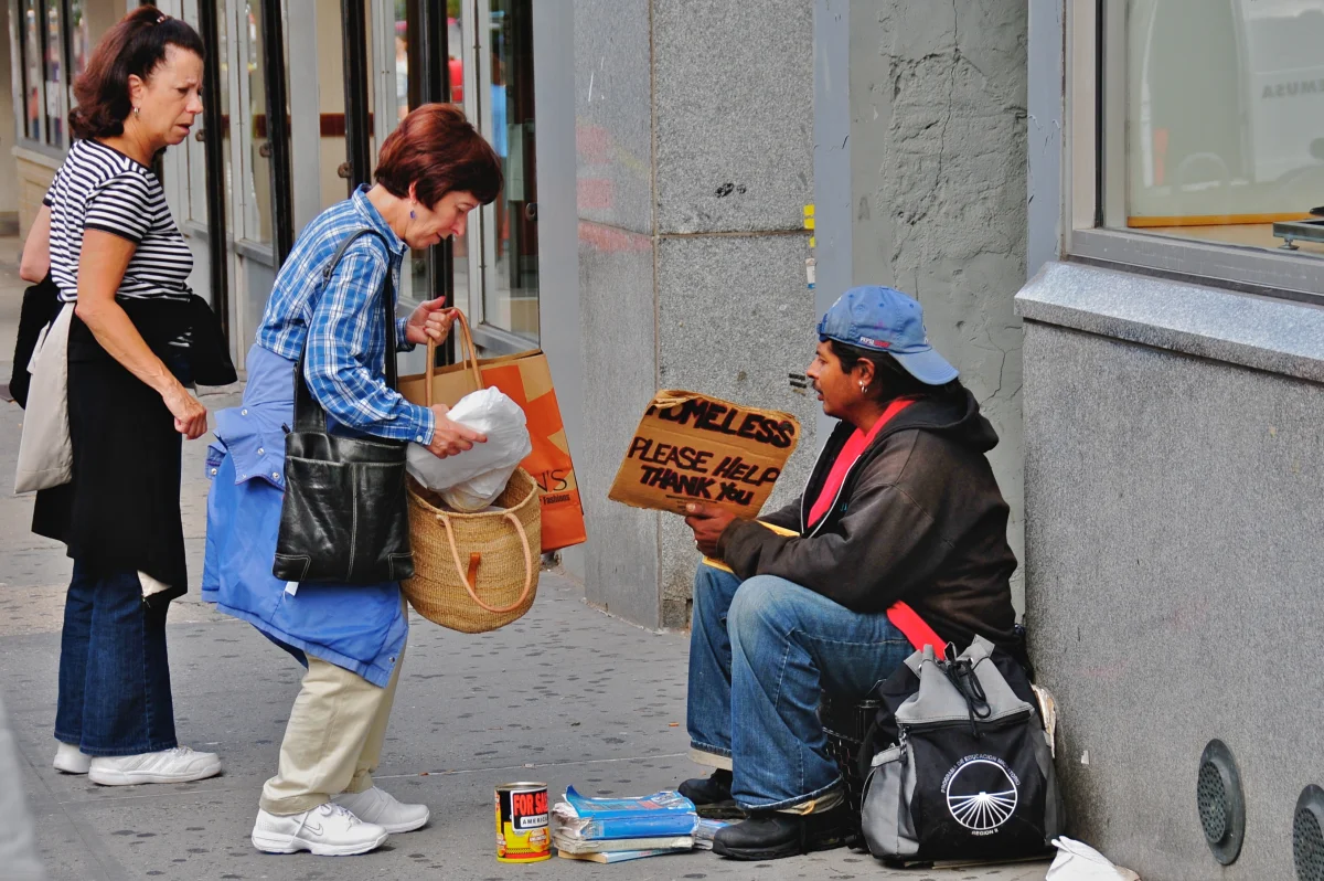 Two passersby on a city sidewalk stop to give a brown paper bag of food to a man sitting against a wall with a cardboard sign asking for help