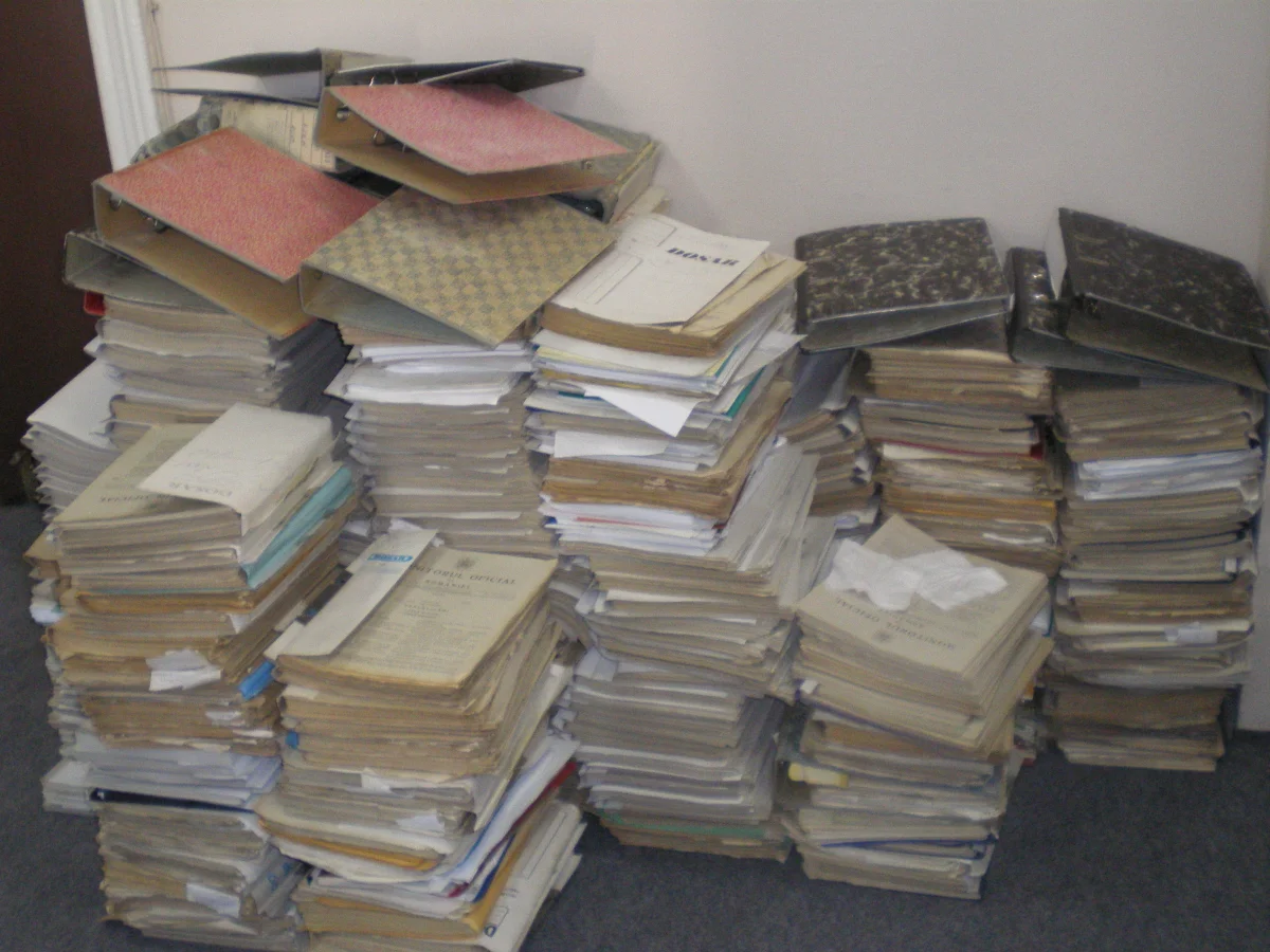 Massive disorganized stacks of bulging cardboard binders and document folders piled against a wall in an office, far taller than knee-height