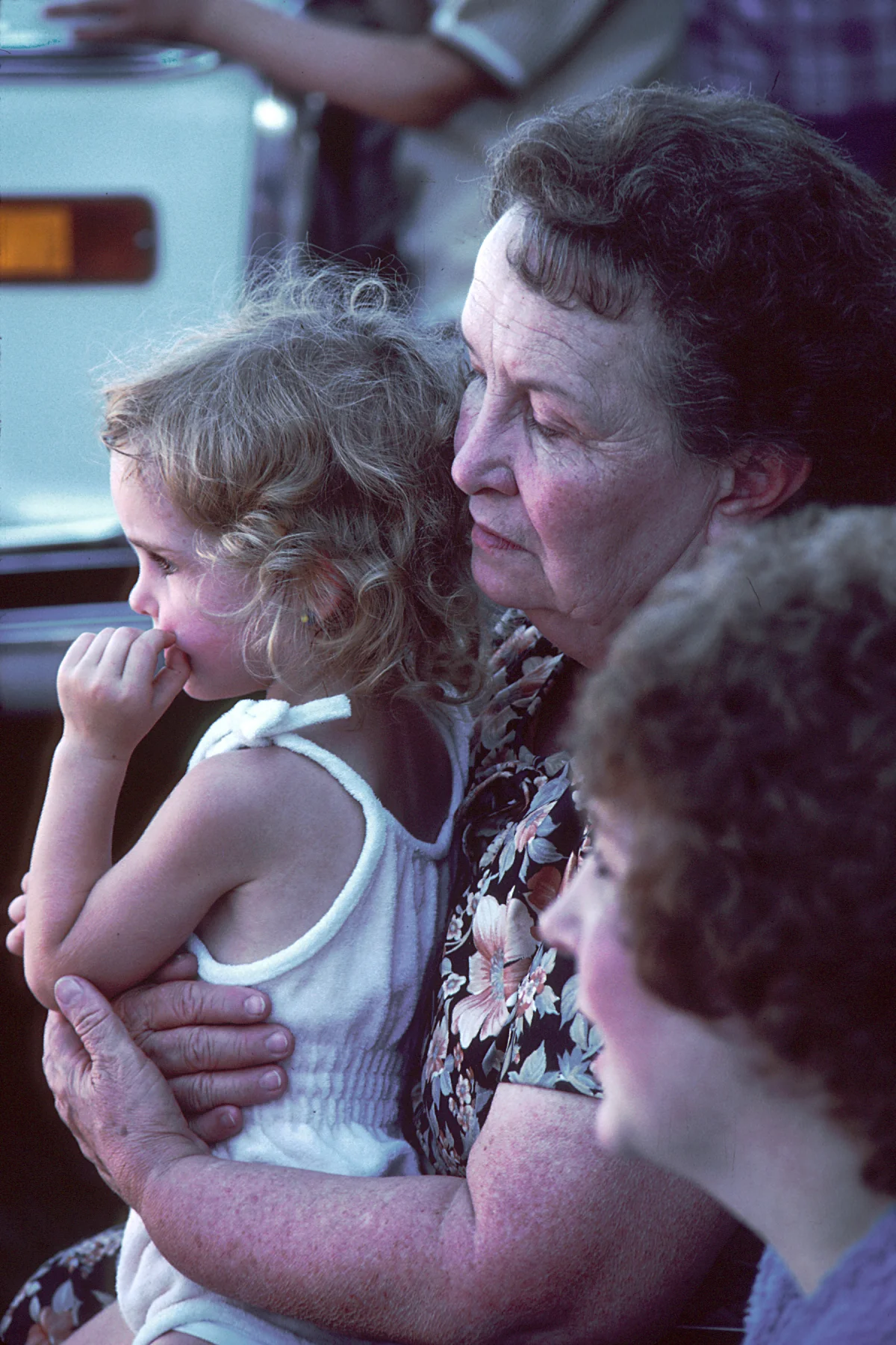 Tender three-generation family scene: an older grandmother holds a small grandchild against her shoulder while another adult sits beside them out of focus