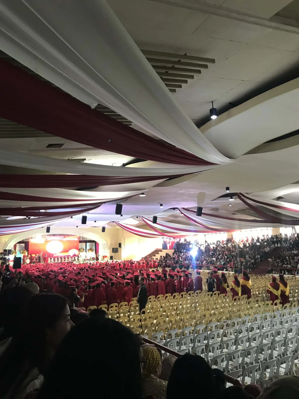 Wide-angle interior view of a large auditorium during a college graduation, hundreds of students in red caps and gowns seated in rows facing a stage decorated with white and red drapes