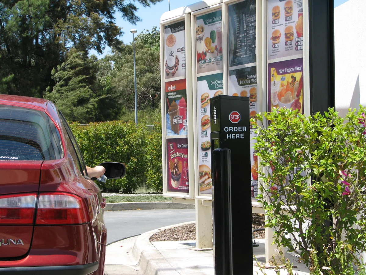 A McDonald's drive-thru lane: a red sedan stopped beside a tall menu board displaying photographs of standardized food items and an 'Order Here' speaker post