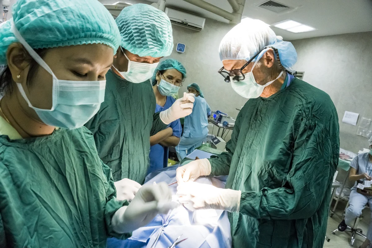 Four members of a surgical team in green scrubs, masks, and head coverings working together over a sterile surgical field in an operating room, one wearing magnifying loupes