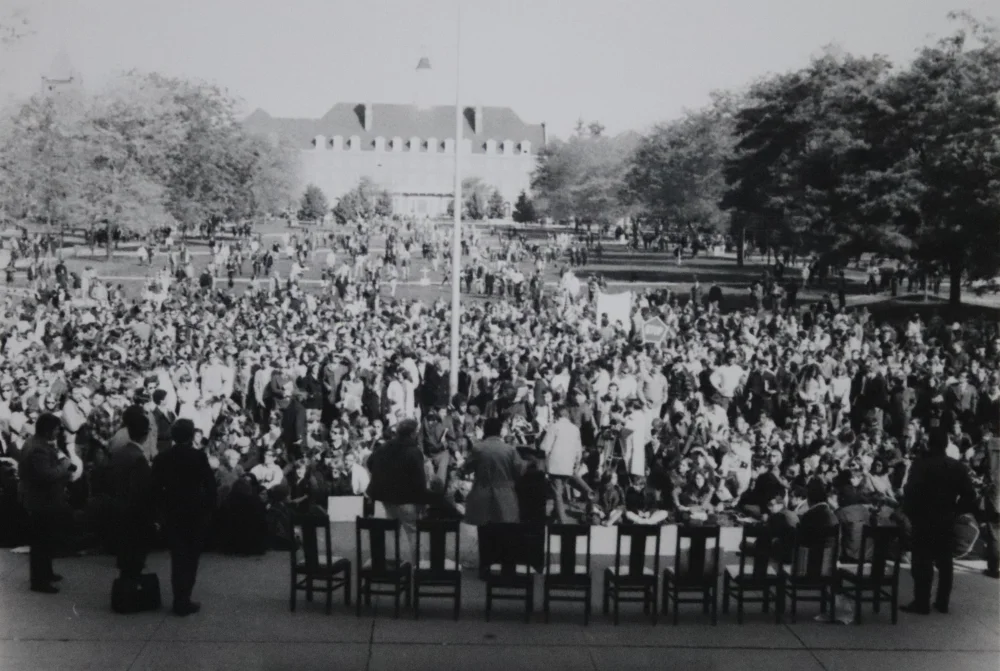 1960s black-and-white photograph of a massive student crowd seated and standing on a university quad facing a stage with empty chairs, with reporters and photographers in the foreground and a campus building in the background