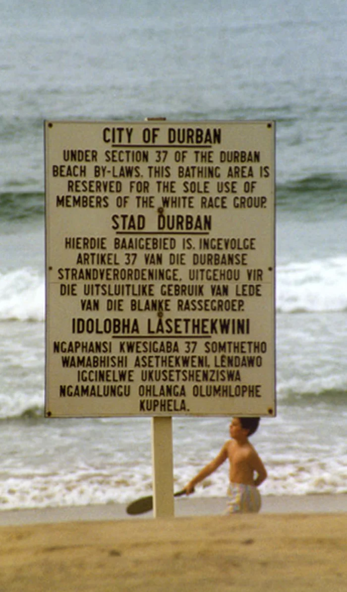 A photograph of a 1989 South African beach sign in three languages reading: 'CITY OF DURBAN: Under section 37 of the Durban Beach By-laws, this bathing area is reserved for the sole use of members of the white race group.' A young child in swimwear stands behind the sign at the water's edge