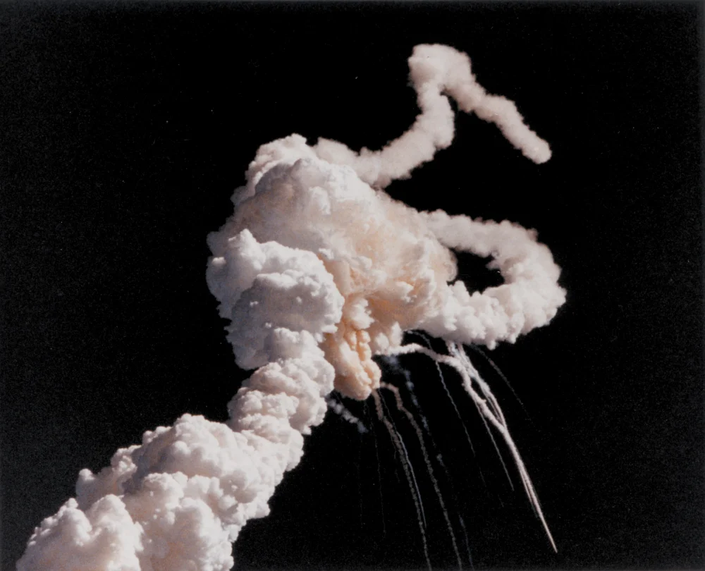 Photograph of the Space Shuttle Challenger explosion on January 28, 1986. A bright white plume of smoke and gas erupts against a black sky, with several smaller debris contrails arcing outward and downward from the central cloud