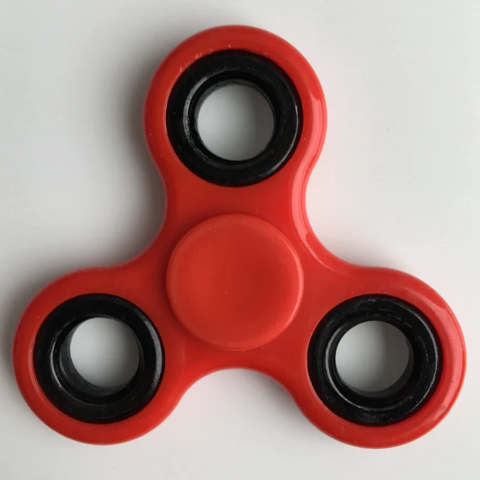 Top-down photograph of a bright red plastic three-arm fidget spinner with black ball-bearing rings at each tip and a central thumb-rest disc, on a white background