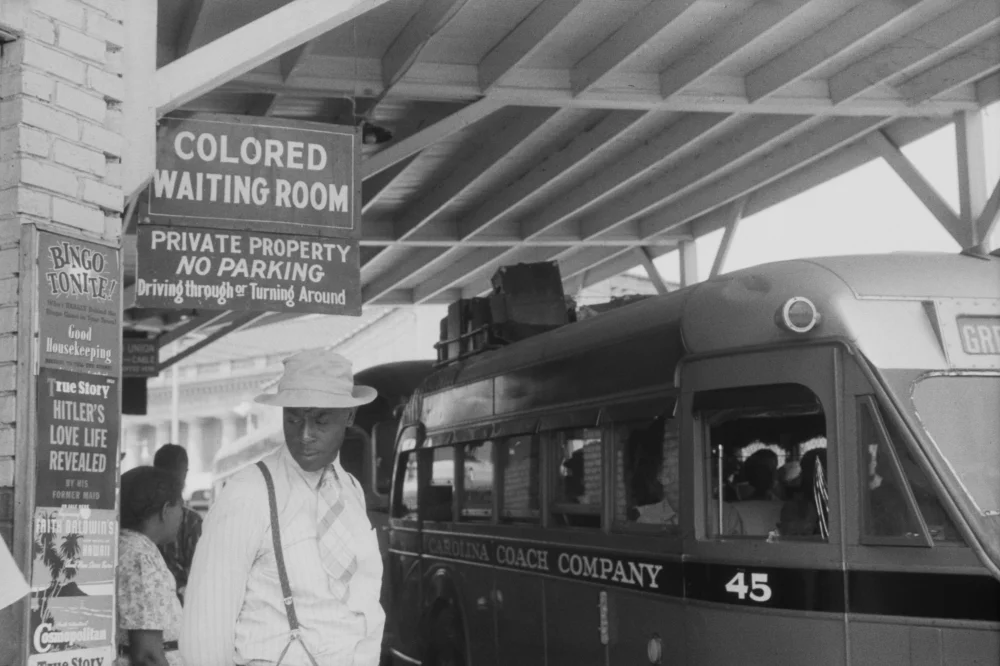 1940 black-and-white photograph of a Black man in a hat and suit standing under a 'COLORED WAITING ROOM' sign at a Jim Crow-era bus station in Durham, North Carolina, while a Carolina Coach Company bus is parked nearby