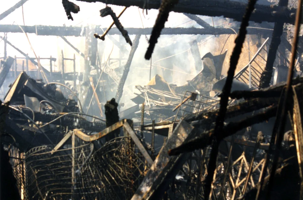 Color photograph showing the burnt-out skeleton of a Los Angeles building after the 1992 riots: charred wooden beams jut at angles, twisted metal grates and rebar lie tangled in the foreground, and pale smoke still drifts through the wreckage