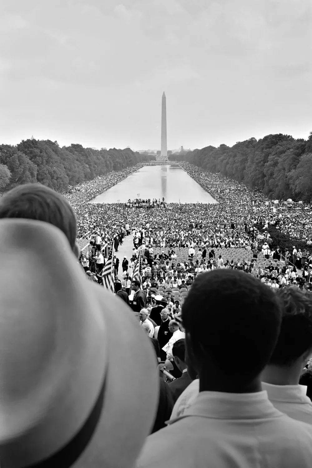 Black-and-white 1963 photograph taken from behind two men in foreground hats, looking out across an enormous crowd filling the National Mall on either side of the Lincoln Memorial Reflecting Pool, with the Washington Monument rising in the distance