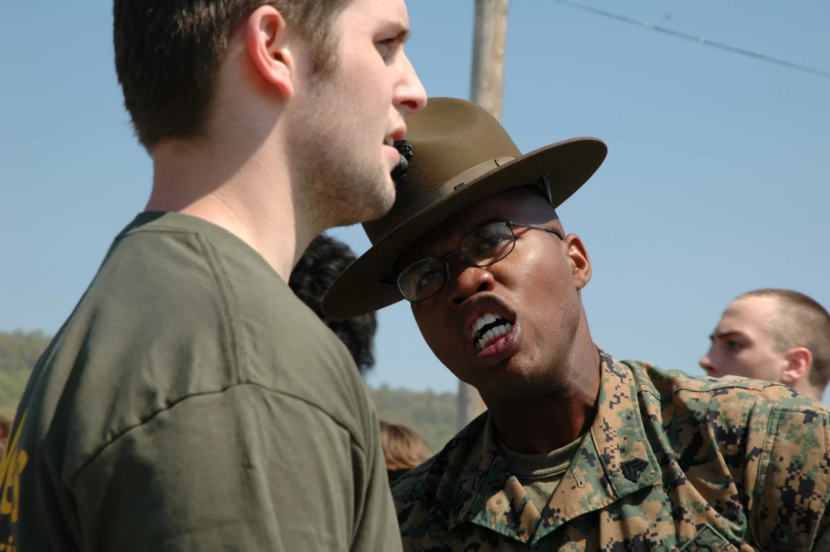 Photograph of a US Marine Corps drill instructor in camouflage utilities and campaign cover running alongside a young civilian recruit on a turf field, supervising physical conditioning before formal enlistment