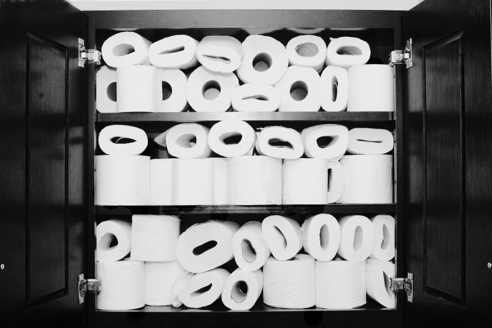Black-and-white photograph of an open kitchen cabinet completely overstuffed with dozens of rolls of toilet paper, packed across three shelves with no remaining space