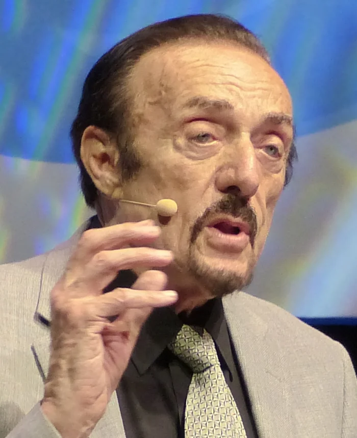 Color portrait of psychologist Philip Zimbardo as an older man with a goatee and dark hair, wearing a gray suit and patterned tie, gesturing animatedly while speaking with a clip-on microphone near his mouth