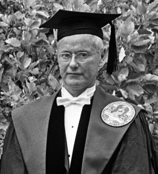 Black-and-white 1965 photograph of sociologist Robert K. Merton, an older man in academic regalia: mortarboard cap with tassel, dark gown over a white bow tie, and a circular medallion pinned to his chest