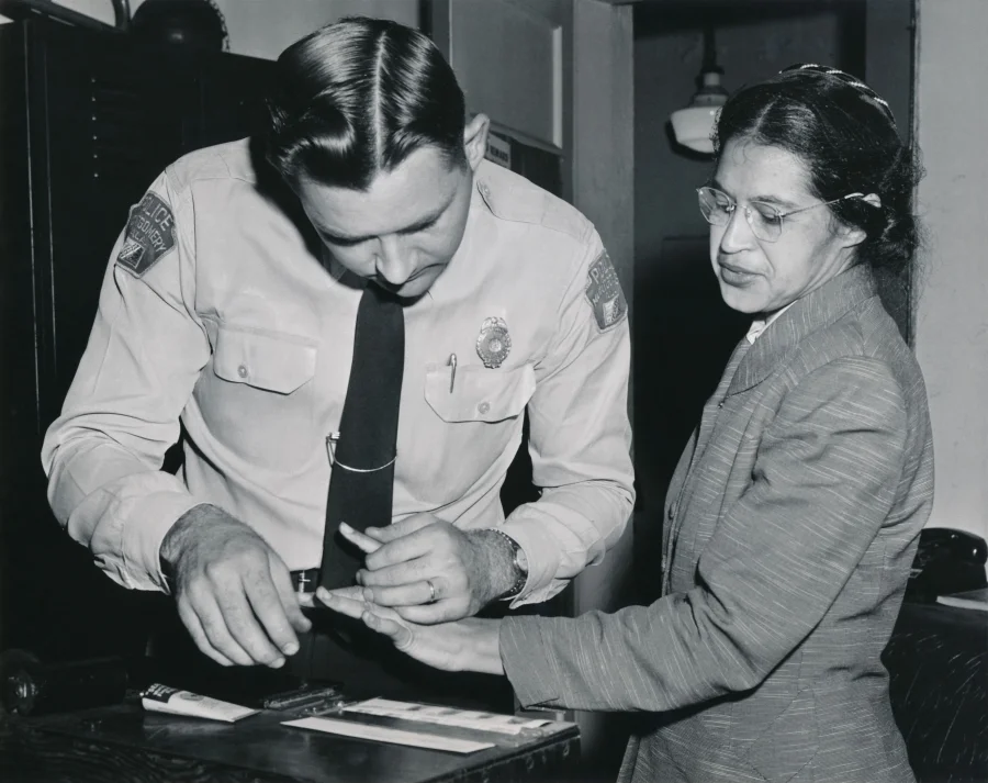 1956 black-and-white photograph of Rosa Parks, a Black woman in a dark suit and glasses, standing next to a uniformed white police officer who is rolling her finger across an ink pad at a station booking desk