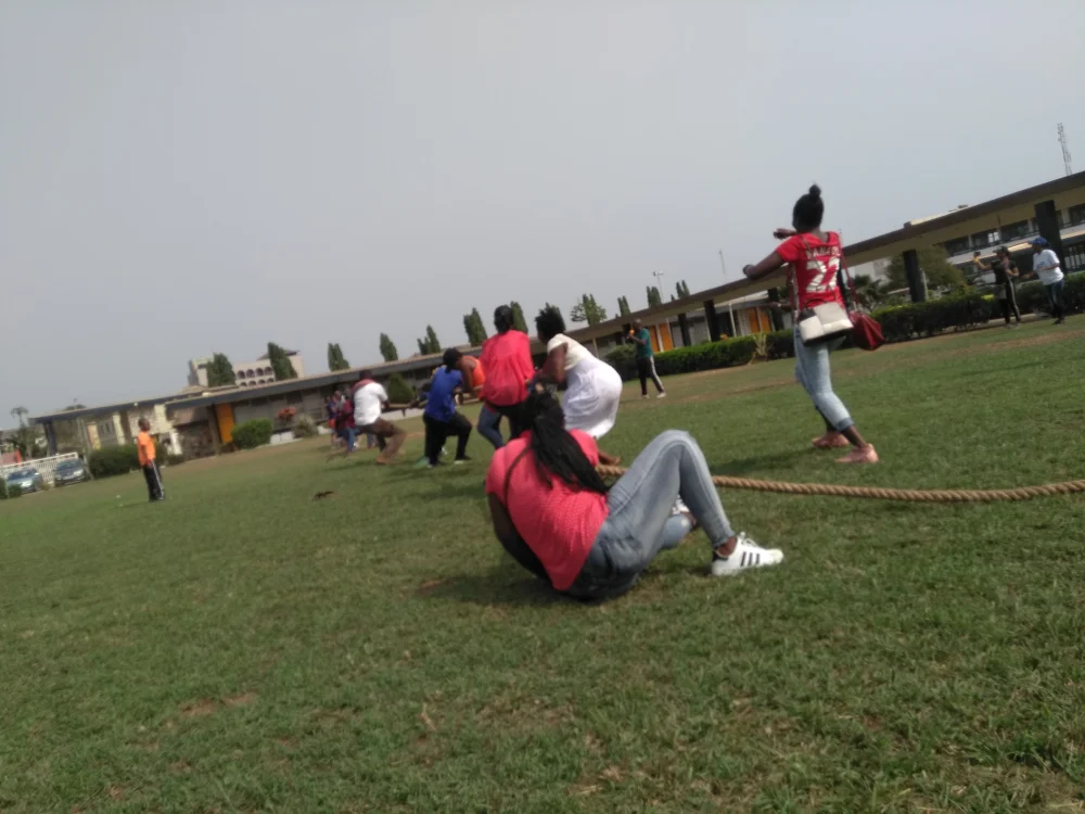 Outdoor photograph of two teams of young adults pulling on opposite ends of a thick rope in a tug-of-war on a grass field. Some pullers are clearly leaning back hard, others appear to be exerting much less force