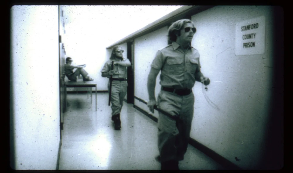 Grainy 1971 photograph from the Stanford Prison Experiment showing two student-guards in khaki uniforms, mirrored aviator sunglasses, and carrying batons walking down a hallway labeled 'Stanford County Prison'. A third guard sits on a table in the background