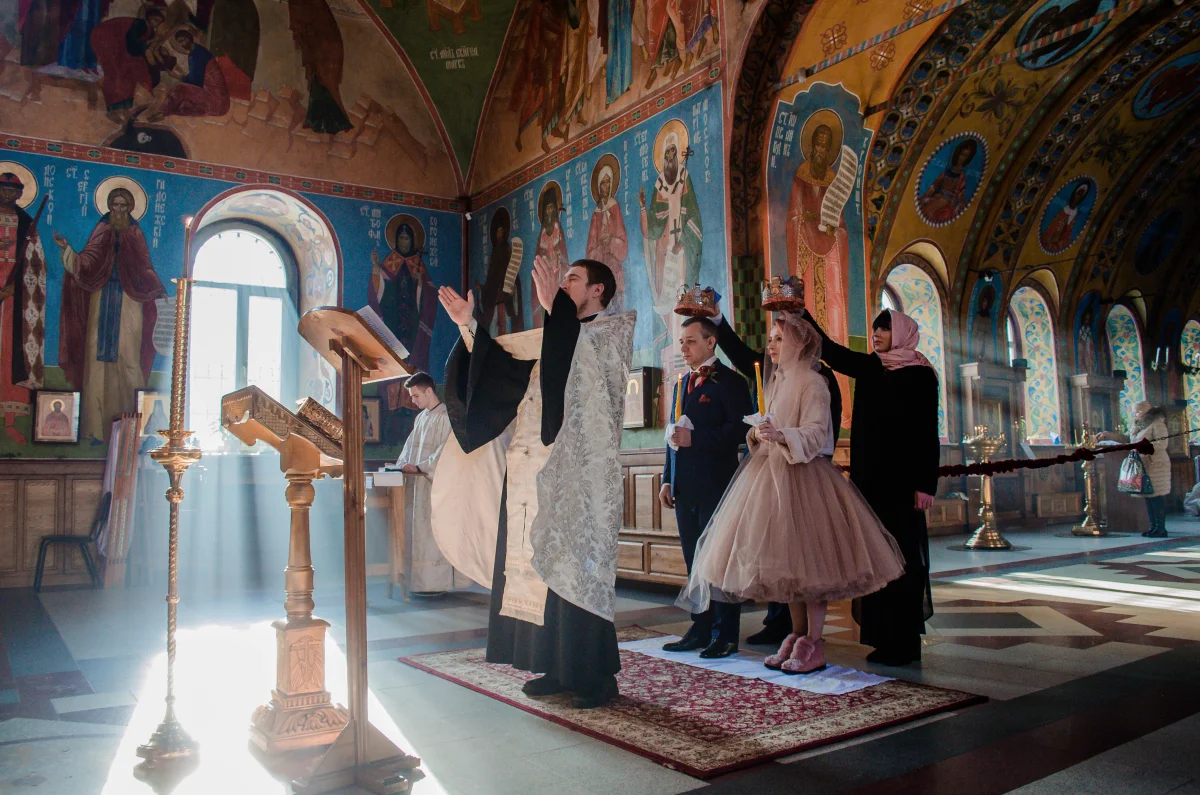 Color photograph of a Russian Orthodox Christian wedding ceremony inside a frescoed church, with a robed priest blessing a bride and groom; sunlight streams through a window onto the floor