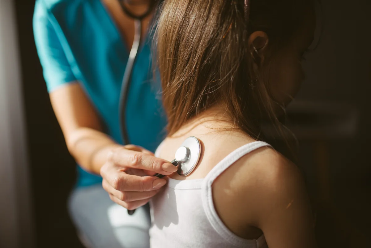 Close-up color photograph of a physician in blue scrubs placing a silver stethoscope chest piece on the back of a young child wearing a white tank top, in a softly lit clinical setting