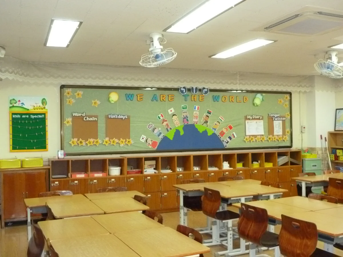 Color photograph of an empty modern elementary school classroom with rows of small wooden desks and chairs facing a corkboard wall covered in student artwork and posters; large windows fill the right side with daylight