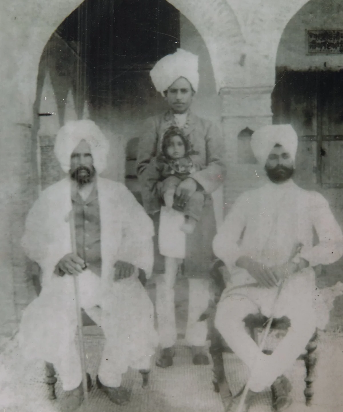 Black-and-white 1909 photograph of three generations of a Sikh family seated together: an elderly bearded man in white robes and turban on the left, a middle-aged man in turban holding a young child in the center, and a younger bearded man in white robes and turban on the right