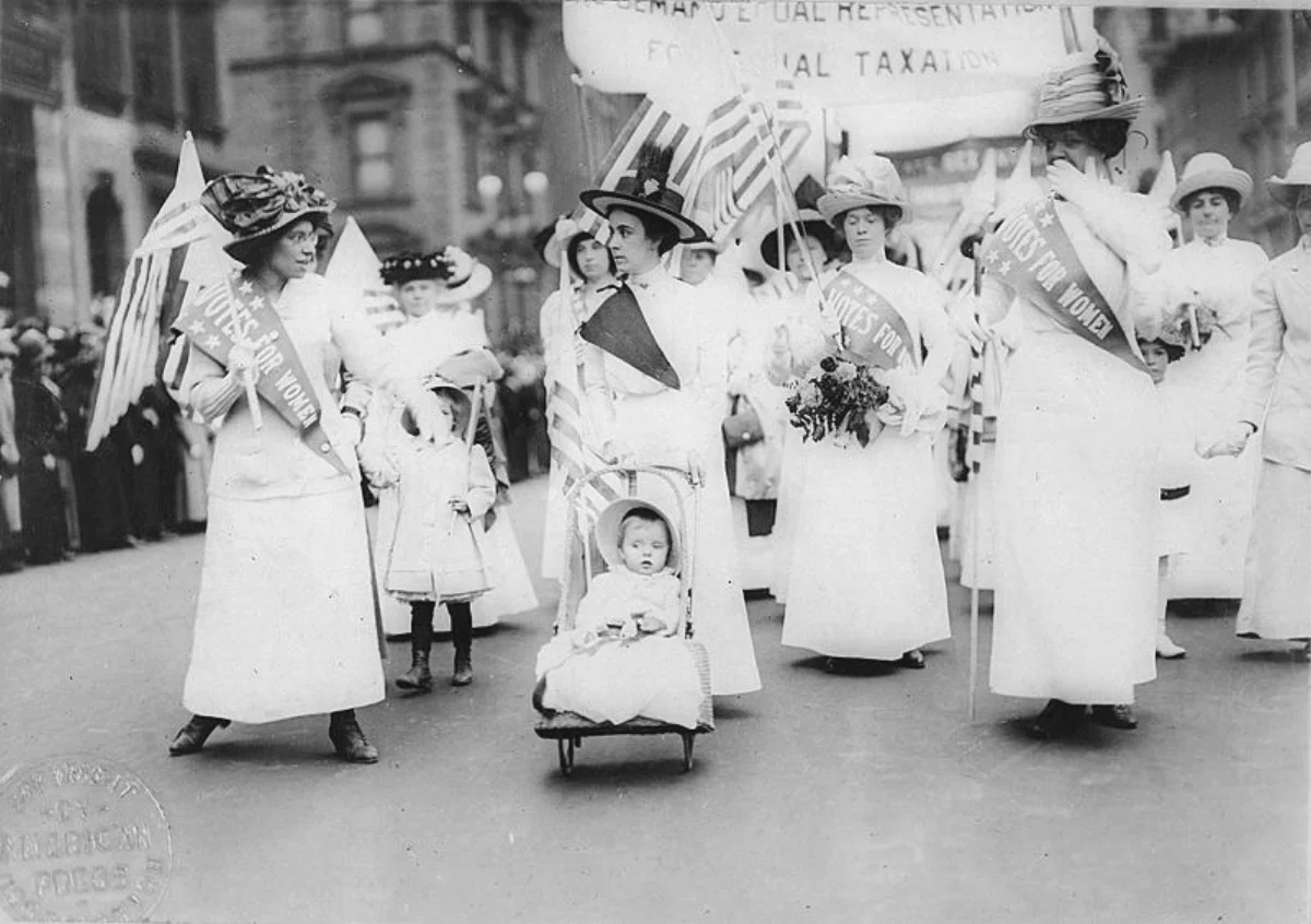 Black-and-white photograph of women suffragists marching in a 1912 parade in New York City, wearing white dresses and 'Votes for Women' sashes, carrying American flags and a banner demanding equal representation