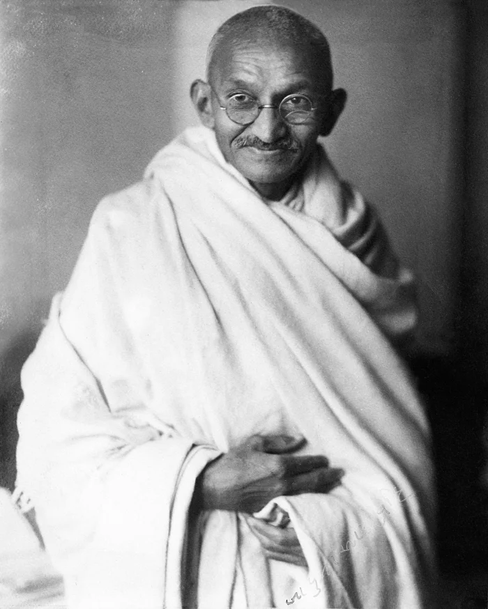 Black-and-white studio portrait from 1931 of Mahatma Gandhi, an older man with a shaved head and round glasses, wrapped in a simple white shawl, smiling slightly at the camera