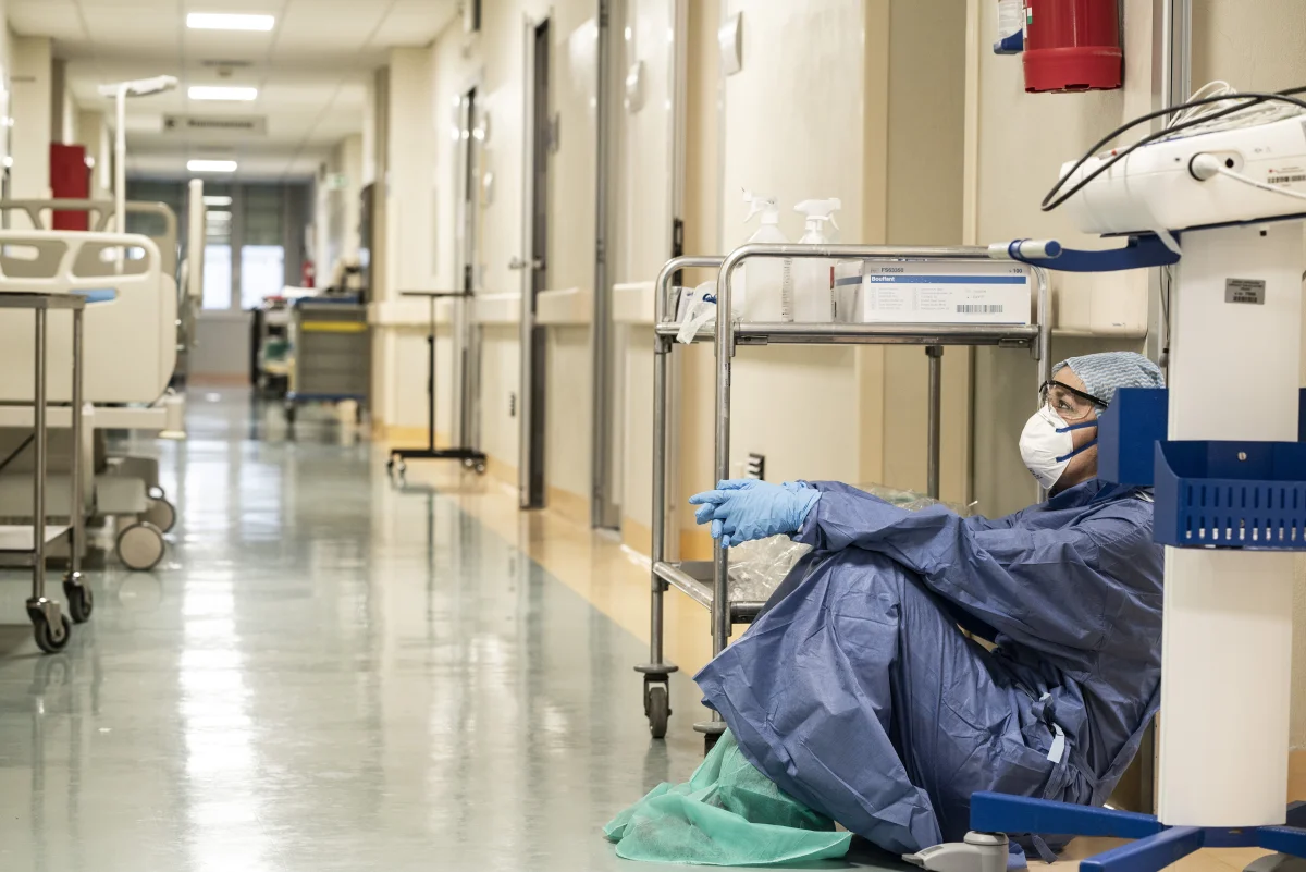 Color photograph of an exhausted healthcare worker in full PPE (gown, mask, gloves, hair cover) sitting on the floor of a hospital corridor next to medical supplies and equipment carts, taking a break during the COVID-19 pandemic