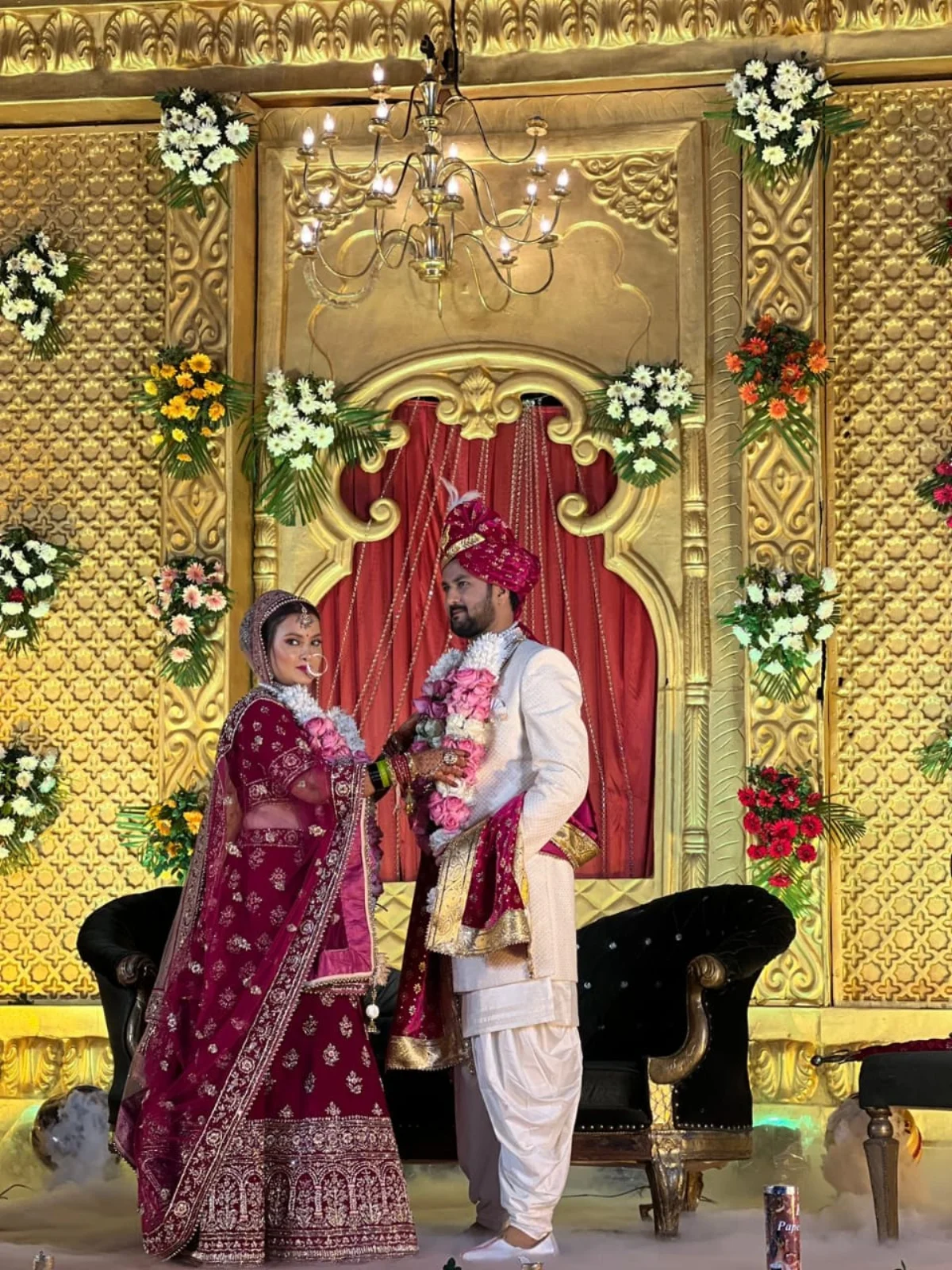 Color photograph of a Hindu bride in a deep red embroidered lehenga and groom in a white sherwani and turban, exchanging garlands while standing on a richly decorated stage with floral arrangements and a chandelier overhead