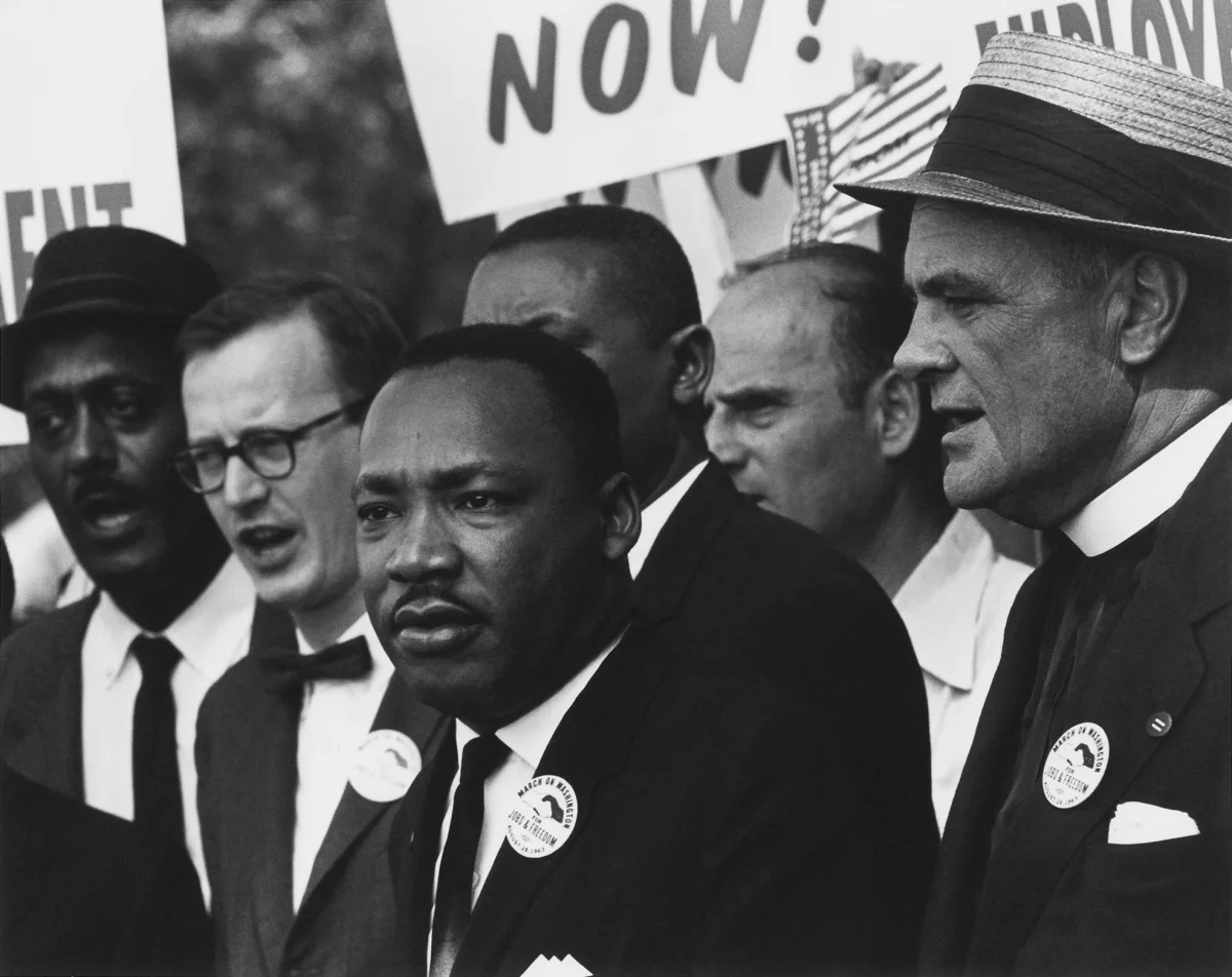 Black-and-white photograph of Dr. Martin Luther King Jr. marching at the front of the 1963 March on Washington, surrounded by other civil rights leaders wearing 'March on Washington for Jobs and Freedom' buttons; protest signs visible behind them