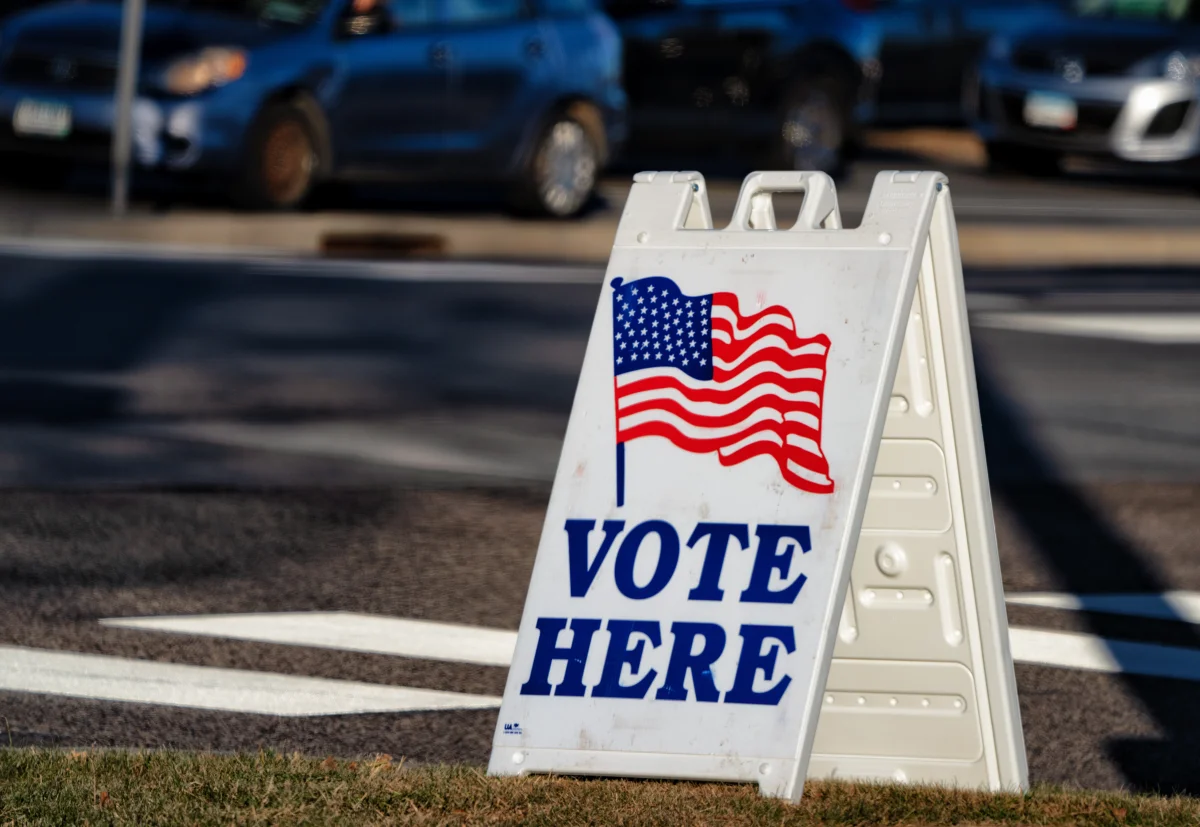 Color photograph of a 'VOTE HERE' sandwich-board sign with an American flag image, set up at the curb in front of a polling place parking lot with cars in the background