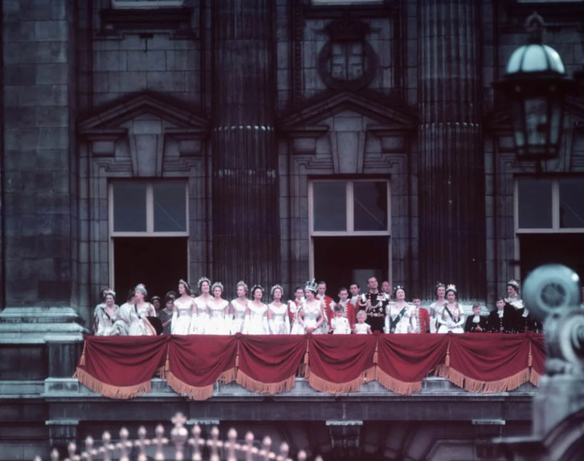 Color photograph of Queen Elizabeth II in coronation robes standing on the balcony of Buckingham Palace in 1953, surrounded by maids of honor in white gowns and members of the royal family in formal regalia behind a red velvet drape