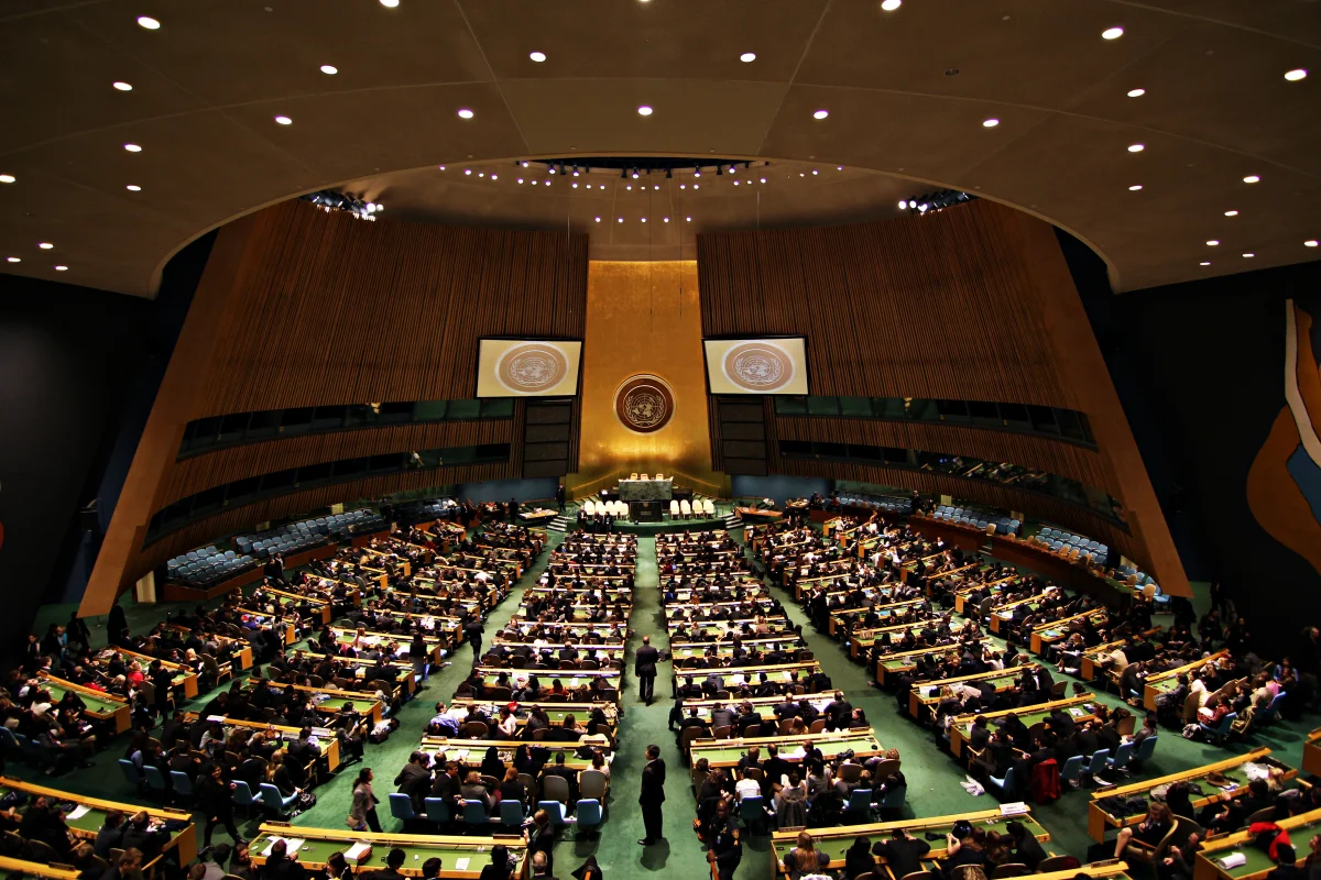 Color photograph of the United Nations General Assembly Hall in New York from the rear gallery, showing rows of country delegations seated below the green marble podium and a large gold UN emblem on the wall