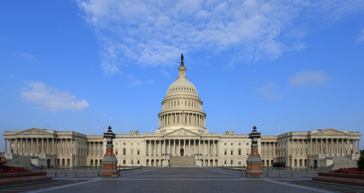Color photograph of the United States Capitol building in Washington DC, viewed straight-on under a blue sky, showing the white dome flanked by the wings of the Senate and House chambers