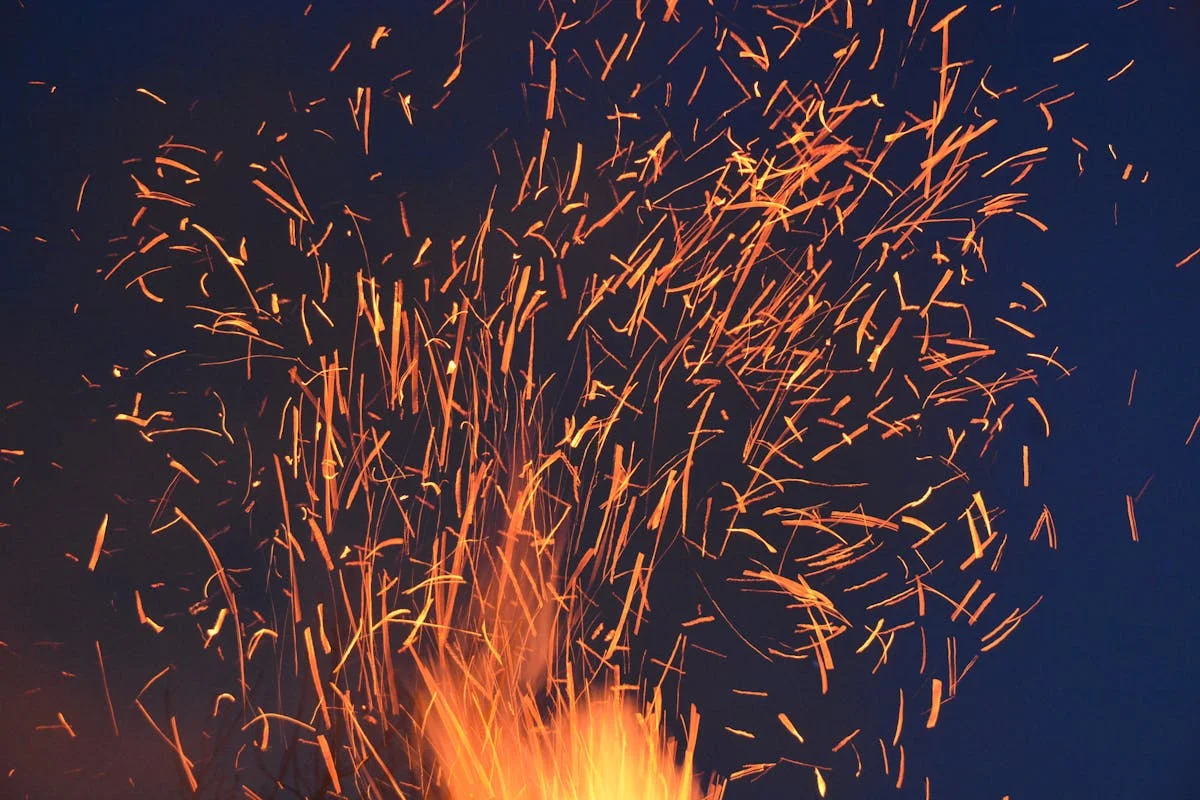 Photograph of a bright fire with orange and yellow flames and sparks flying upward against a dark background
